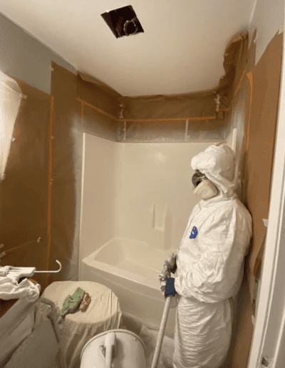 Person in protective gear stands in a bathroom under renovation, with walls covered in plastic sheets and tools scattered around. Ceiling hole visible; appears to be involved in remediation or cleaning work.