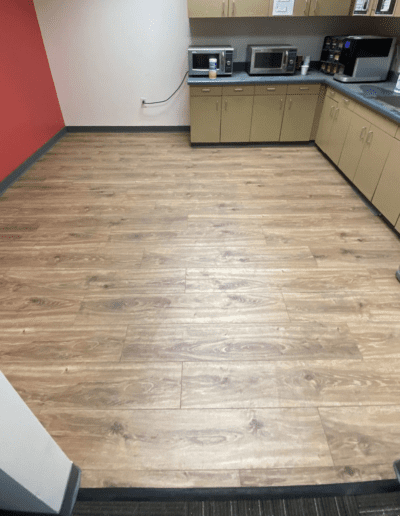 A kitchen with wooden floors, beige cabinets, and stainless steel appliances. The counter has a microwave, coffee machines, and storage containers. The walls are red and white.