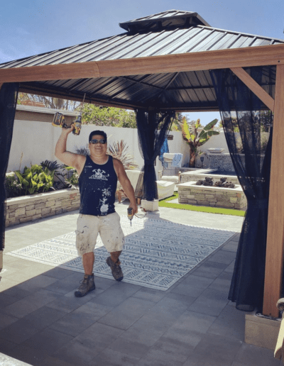 A person in a sleeveless shirt, shorts, and work boots holds a drill and poses under a gazebo structure with a pool in the background.