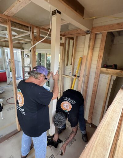 Two construction workers install a wooden beam in a partially framed room, with one holding the beam and the other securing it with a tool.