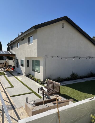 A two-story house with white walls and a dark roof has a backyard featuring a grassy area, a wooden bench, and a pergola. String lights are hung above the yard, and a road is visible on the side.