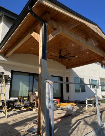 Two workers are renovating a house with a newly constructed wooden patio covering and unfinished railing. Various tools and materials are scattered around.