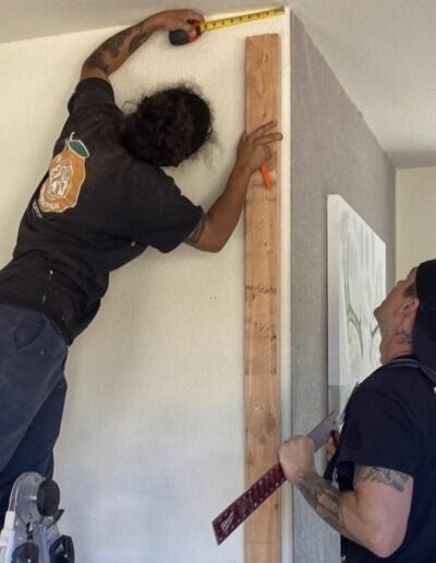 Two individuals performing home renovations; one person is measuring the wall height with a tape measure and the other is holding a wooden plank and a level.