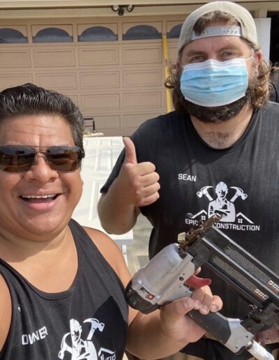 Two men wearing "Epic Construction" shirts pose in front of a garage door; one holds a nail gun and the other gives a thumbs up. The man on the right is wearing a surgical mask.