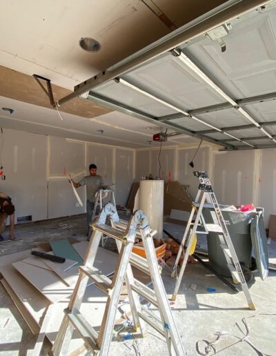 Two individuals work on home improvement in a garage, which is filled with ladders, tools, and construction materials. The garage door is open, and the scene is brightly lit.