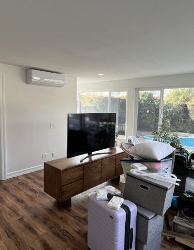 A room with wooden flooring containing a TV on a stand, stacked boxes, a potted plant, and a view of an outdoor pool through large windows. The room appears to be in disarray.