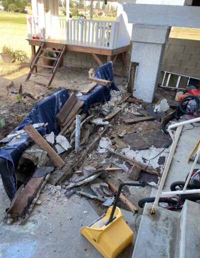 A damaged outdoor area featuring rubble, broken wood, concrete debris, and tools. A large hole is visible in the ground. A playhouse with a small porch stands in the background.