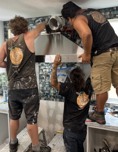 Three workers are installing a kitchen range hood, with one individual pouring a substance through the top while the others hold and secure the hood in place.