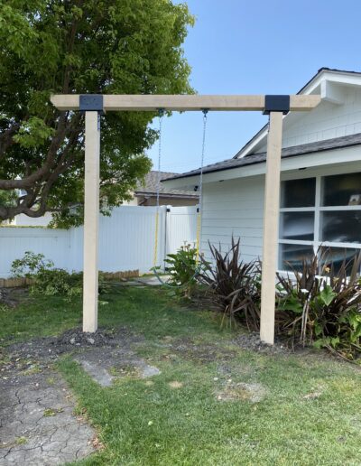 A backyard swing set with a wooden frame and two swings hangs in front of a white house. A tree and plants are in the background.