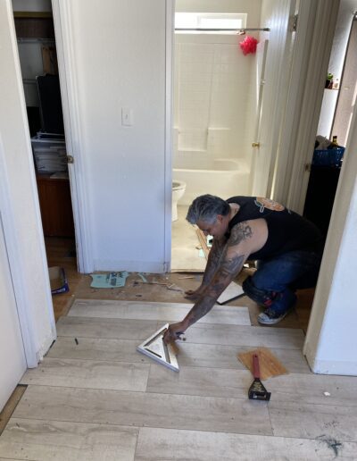 A man wearing a black tank top with tattoos on his arms is installing wood flooring in a hallway near a bathroom. Tools and materials are scattered around.
