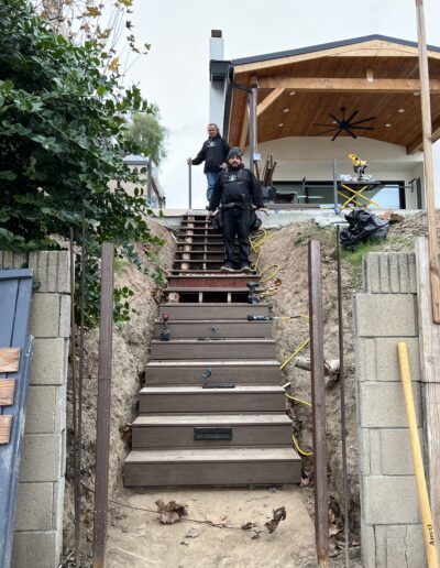 Two workers stand on wooden steps under construction leading up a slope to a house. Construction tools, cables, and materials are scattered around.