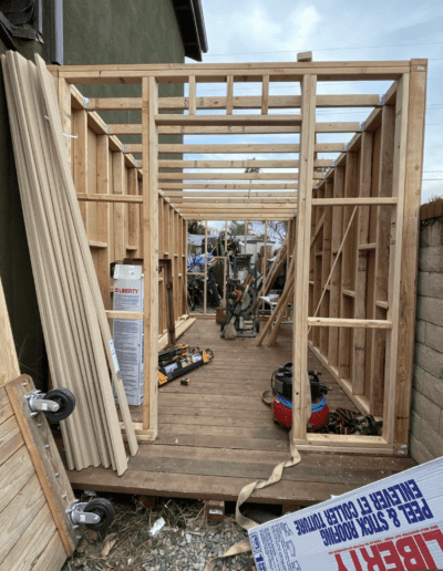 A partially constructed wooden frame structure with various tools and building materials scattered on the floor and nearby. A brick wall and neighboring building are visible in the background.