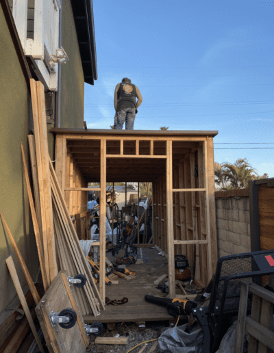A person in a helmet and construction vest stands on the roof of a partially built wooden shed alongside various tools and materials.