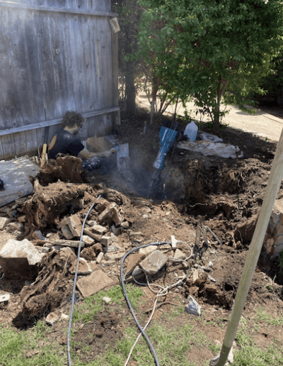 A person is repairing a large hole in the ground next to a wooden fence, surrounded by various tools and pieces of equipment.