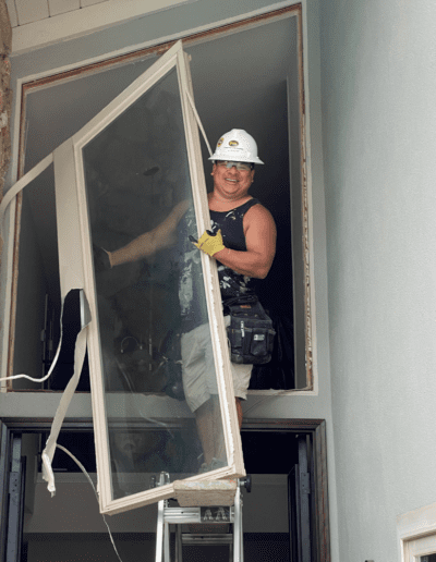 A construction worker wearing a hard hat stands on a ladder, holding a large window frame. He is smiling and positioned in an open window space of a building.