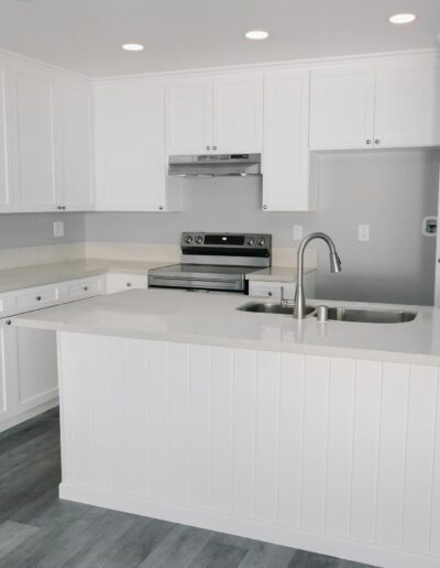 Modern kitchen with white cabinets, stainless steel appliances, and gray countertops. The room features a central island with a sink and wood-paneled accents. The floor is gray wood.