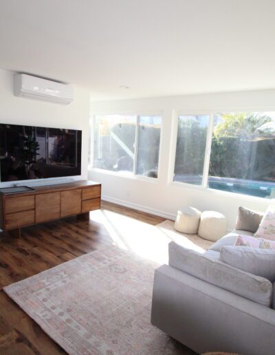 Bright living room with large windows, a gray sofa, a flat-screen TV on a wooden stand, a rug, some poufs, and a potted plant. Wood flooring and storage area visible in the background.