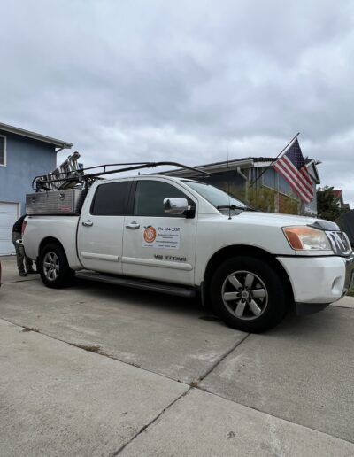 White pickup truck with a ladder rack, parked in a residential driveway. The truck has a business logo and contact information on the door. An American flag is visible in the background.