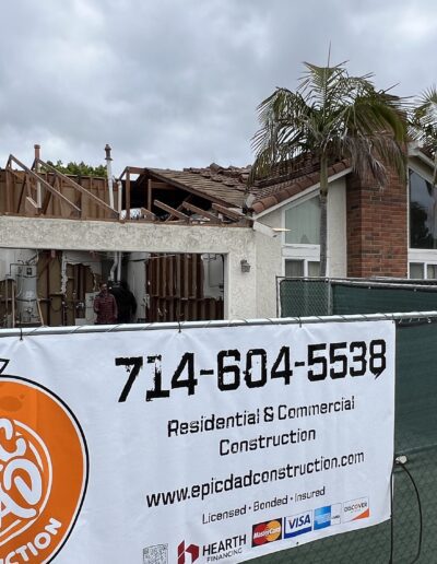 Workers are seen on the roof and interior of a house undergoing construction, behind a green fence with a banner displaying "Epic Dad Construction" and contact details.