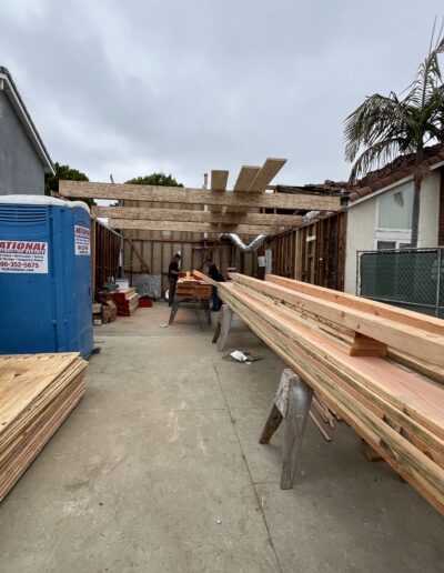 A construction site with wooden beams and panels being assembled, a portable toilet on the left, and a worker in the background with a saw and materials spread around.