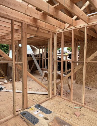 A house under construction with wooden framing and exposed beams. Various construction tools and materials are scattered throughout the site. Roof tiles and trees are visible in the background.