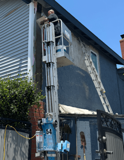 A person stands in a lift working on the exterior wall of a house, which has a partially completed gray finish. A ladder leans against the house and construction tools are visible below.
